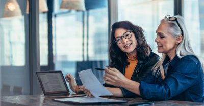 Two female professionals at a desk with a laptop reviewing some paperwork